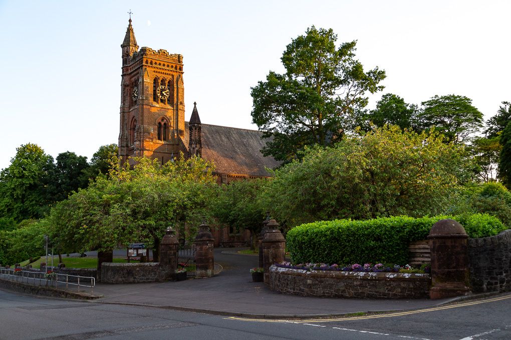 Moffat's Historic stone church with a tall bell tower stands behind hedges and a stone wall at sunset in a rural setting.
