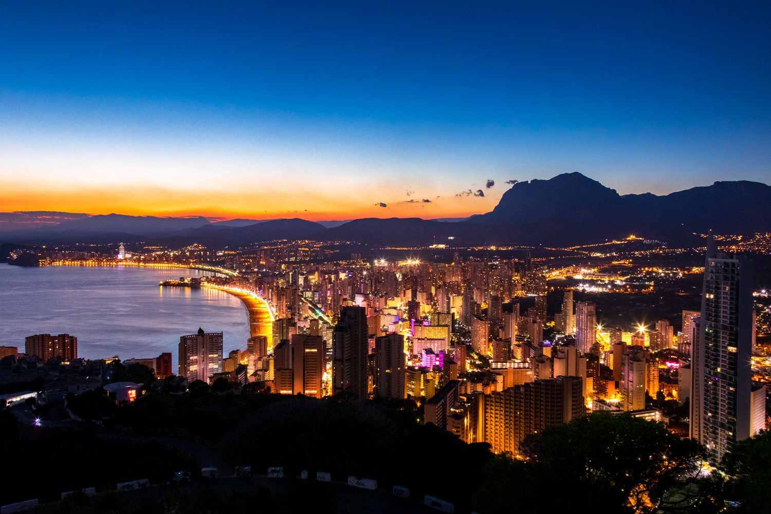 Stunning aerial view of Benidorm city skyline at dusk with glowing lights along Mediterranean coastline and mountains.