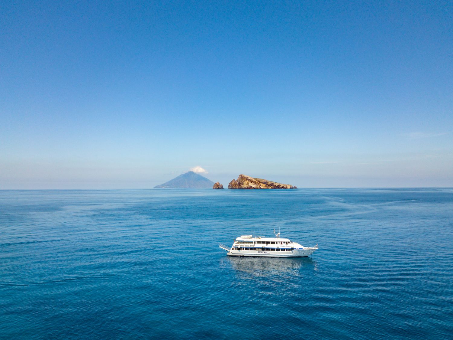 A white passenger ferry cruises on calm turquoise Mediterranean waters near volcanic Stromboli Island on a clear day.