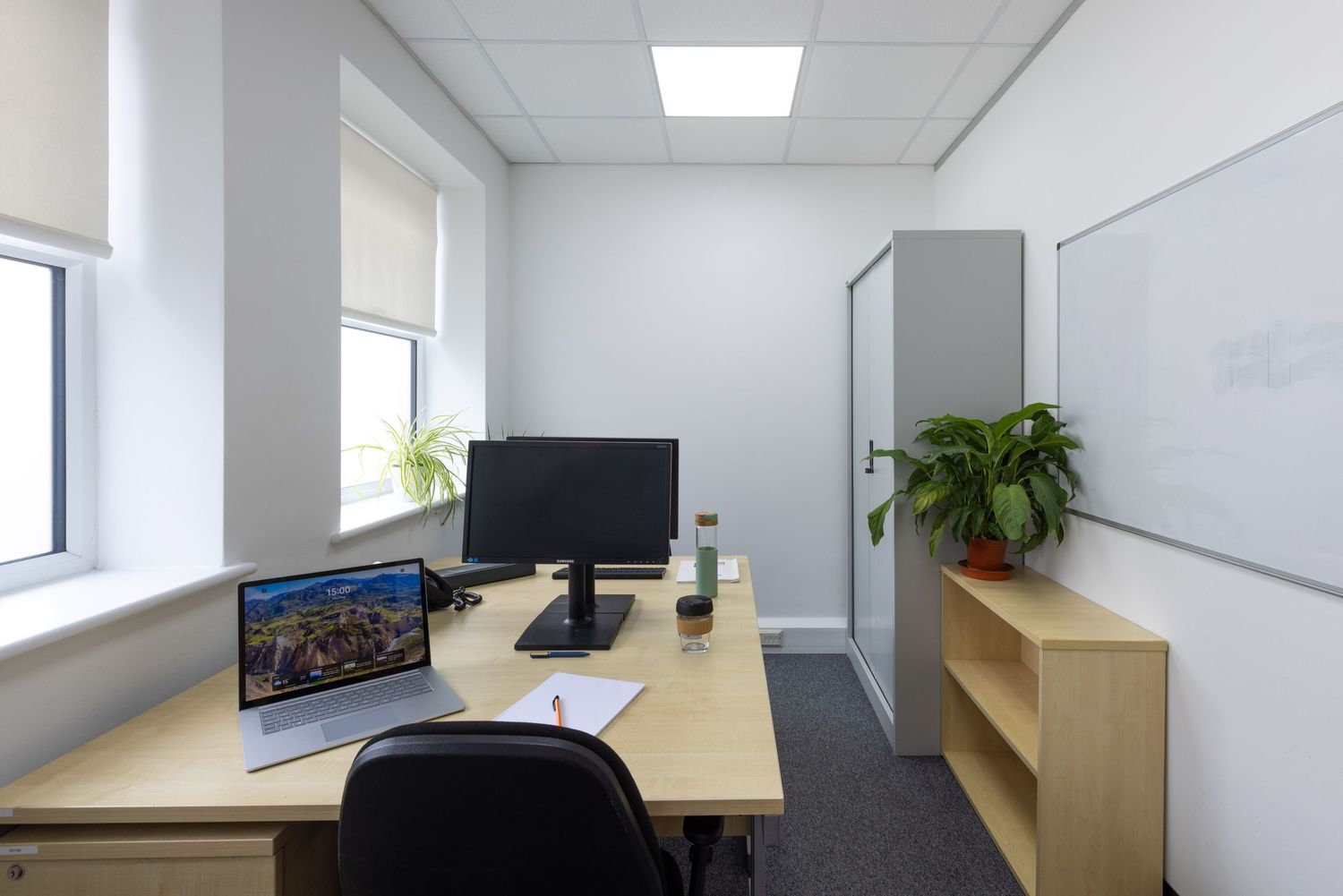 Modern small office with wooden desk, computer monitor, laptop, bookshelf, whiteboard, and green plant by bright window.