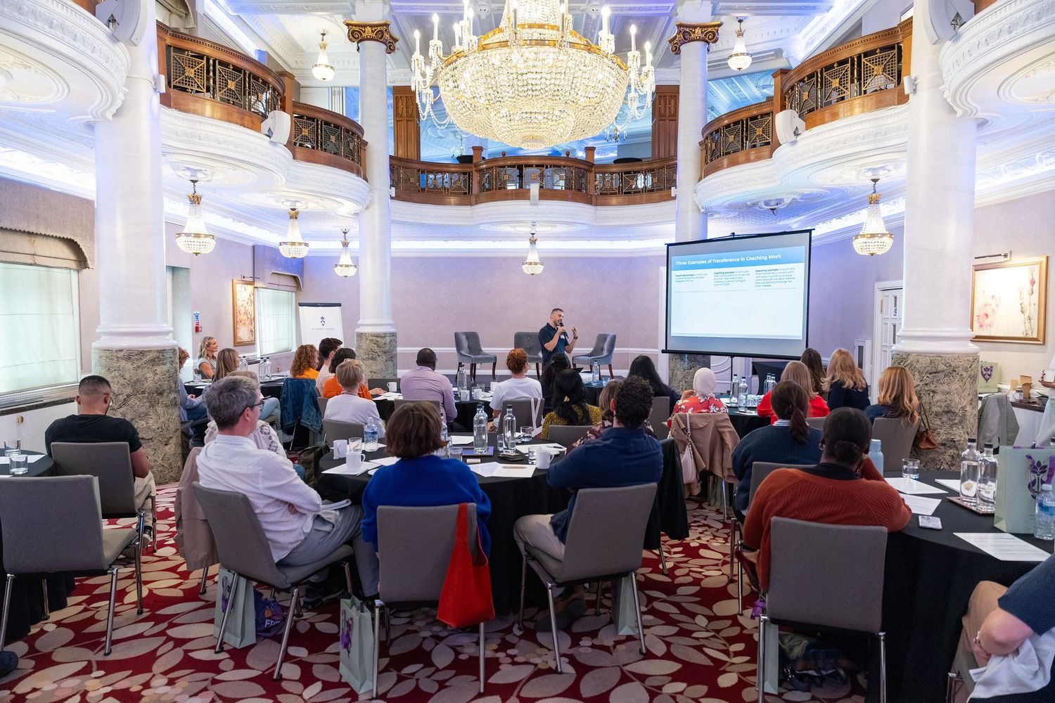 Attendees listen to a presenter at a corporate conference in an elegant ballroom with chandeliers and a projection screen.
