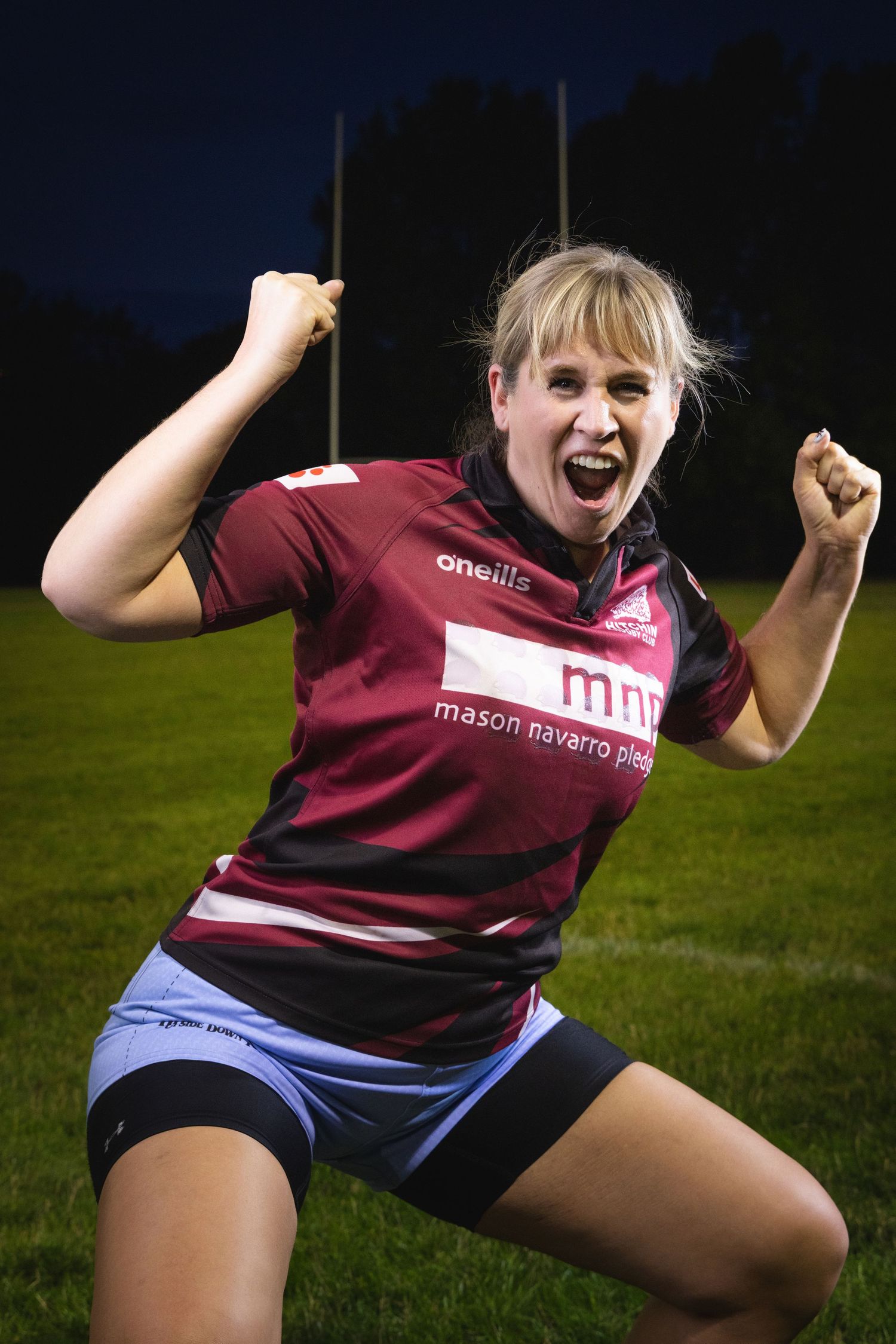 Excited female rugby player in maroon Mason Navarro Pledge jersey celebrating victory on green field at night.