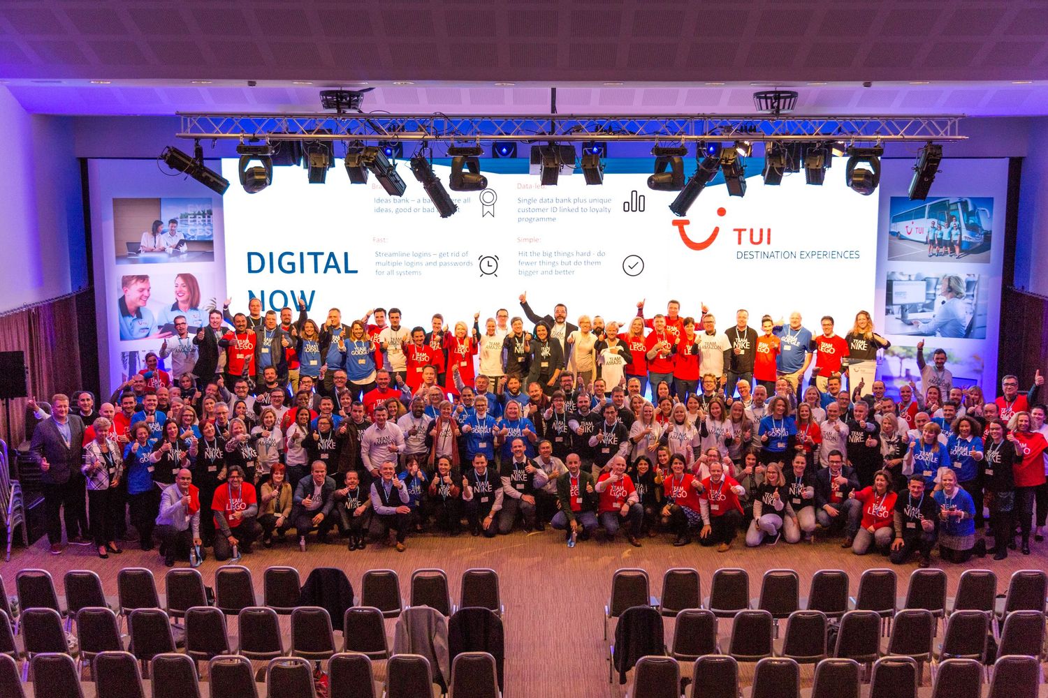 Large group photo at TUI Digital Now conference, attendees in colorful attire posing on stage with branded backdrop.