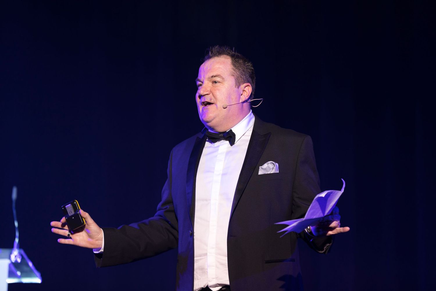 Man in tuxedo performing on stage, gesturing expressively while holding papers under blue spotlight.