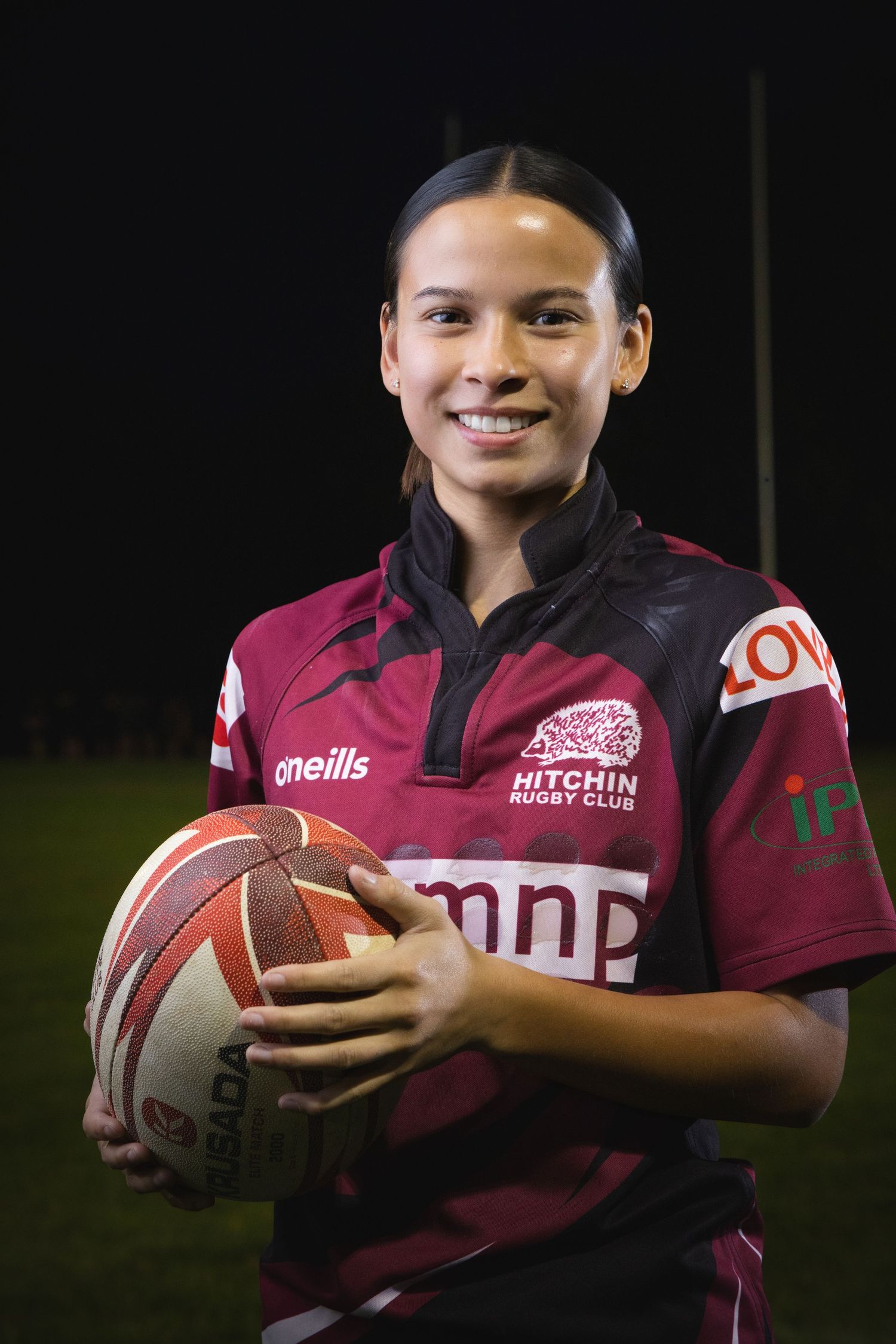 Young woman in Hitchin Rugby Club jersey smiling while holding a rugby ball on a field at night.