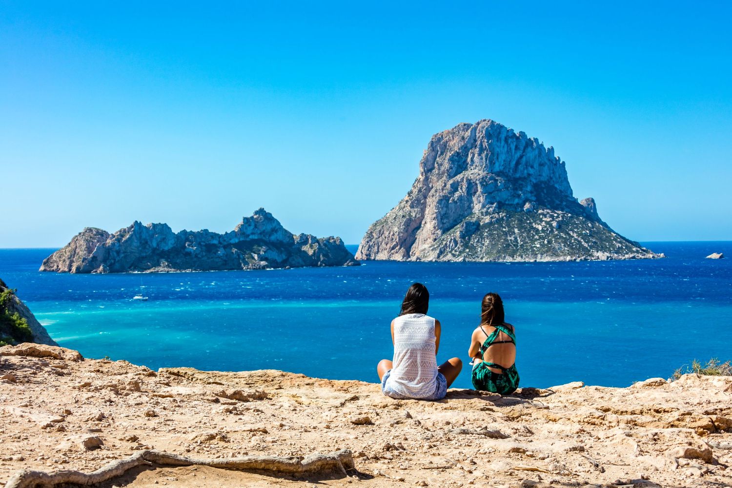 Two women sitting on rocky cliffs overlooking the stunning turquoise waters and Es Vedra island in Ibiza, Spain.