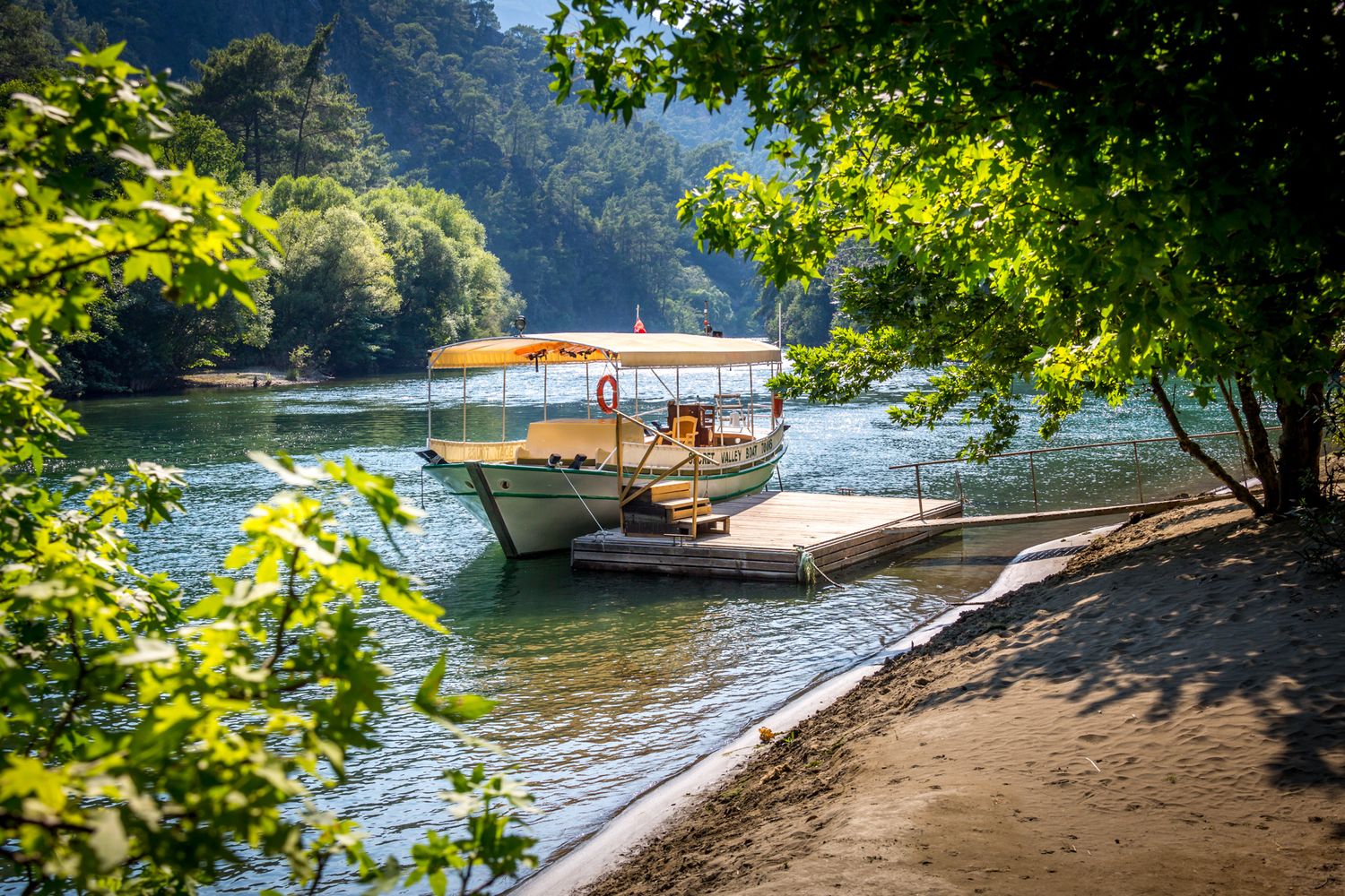 A wooden tourist boat docked along a scenic riverbank surrounded by lush green trees and foliage.