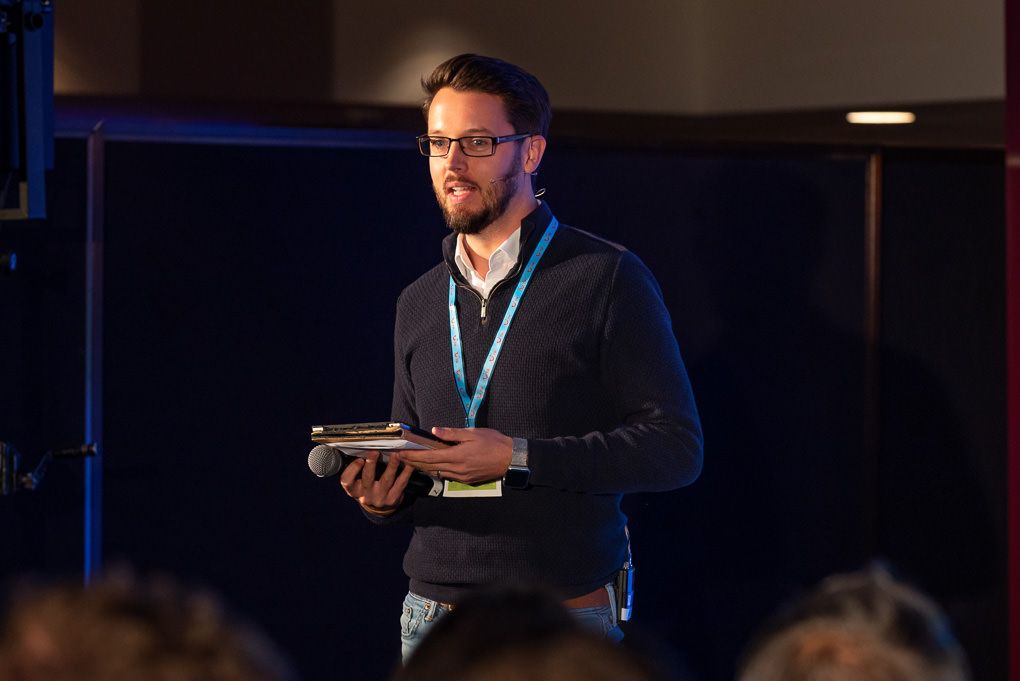 A speaker presents at a conference podium with blue stage lighting in the background. Conference photography at The Brewery in London.