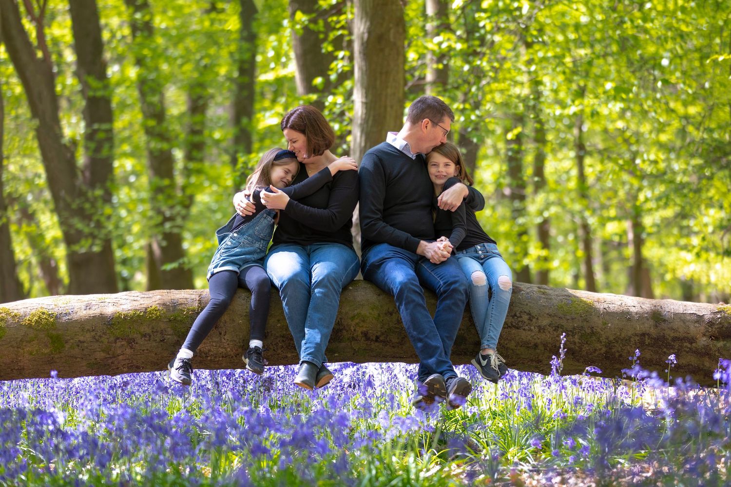 Family of four sitting on a fallen log in a bluebell woodland, sharing tender embraces surrounded by purple flowers.