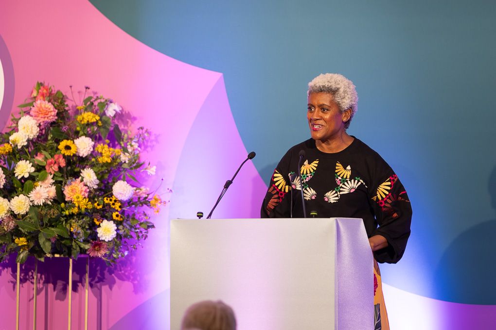 A speaker stands at podium with colourful floral arrangements and dramatic pink and blue stage lighting for the RHS Britain in Bloom Awards