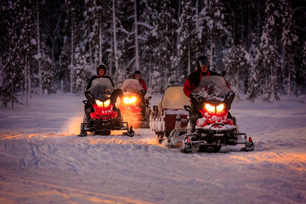 Snowmobiles with glowing headlights traverse through a snowy forest landscape at dusk creating a magical winter scene in Lapland, Finland.