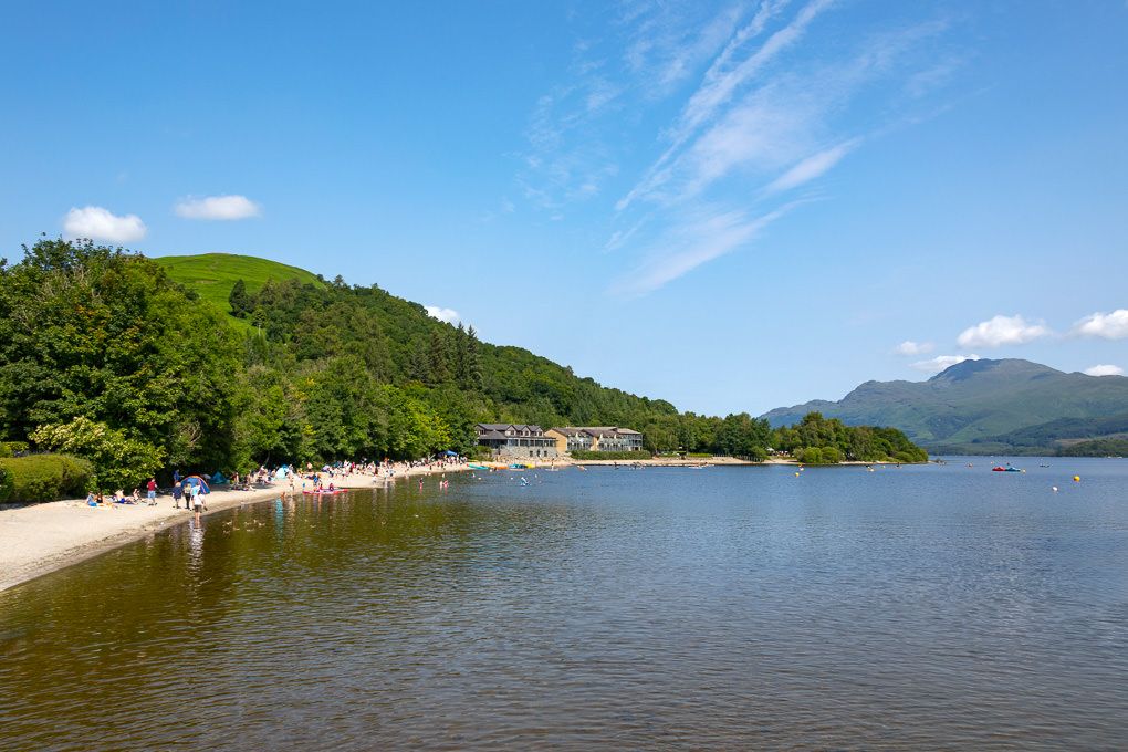 Panoramic view of a Loch Lomond surrounded by lush green mountains and forests under a bright blue sky.