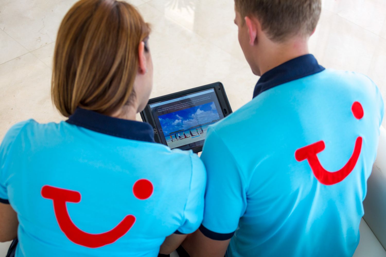 Two TUI employees wearing branded blue shirts complete with red TUI Smile, looking at a tablet computer together in an office.