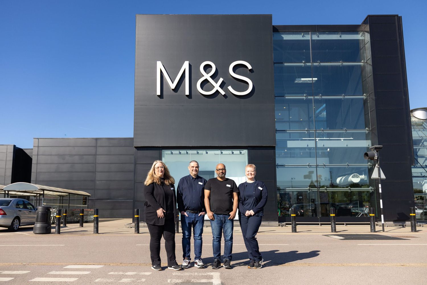 Four M&S employees standing outside a modern Marks & Spencer store entrance on a sunny day.