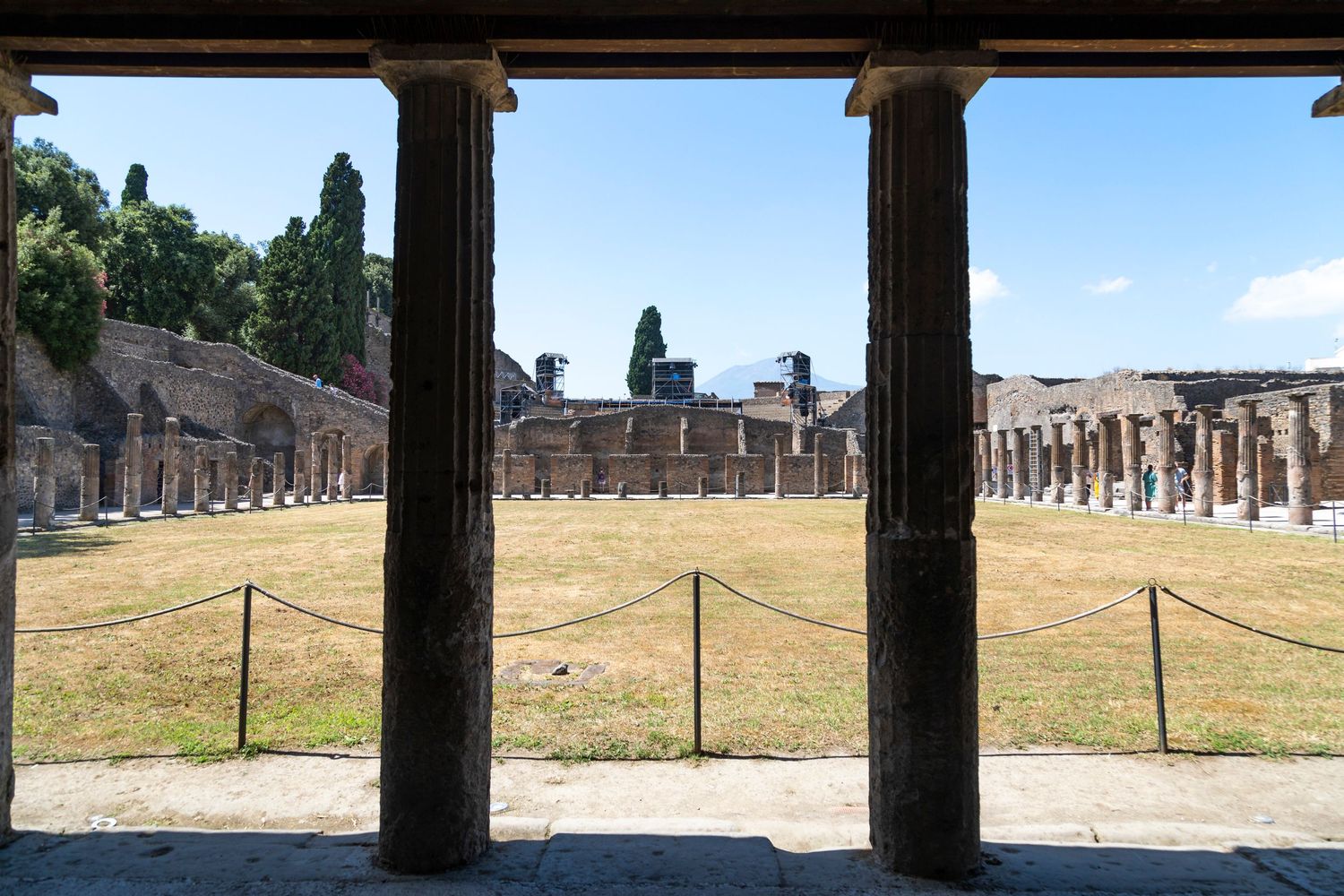 Ancient Roman ruins viewed between two stone columns, showing a grassy courtyard enclosed by rope barriers and a colonnade under a blue sky.