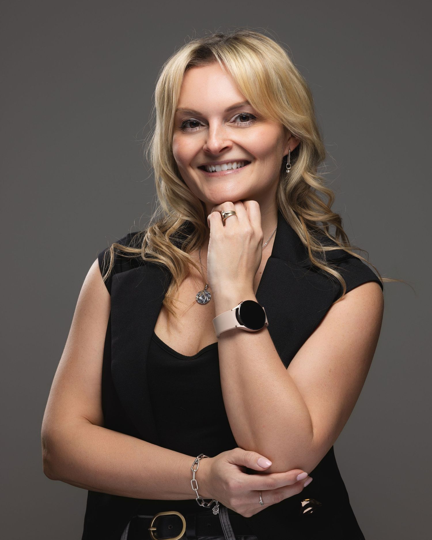 Professional woman in black outfit smiling confidently, wearing a smartwatch and bracelet, against gray background.