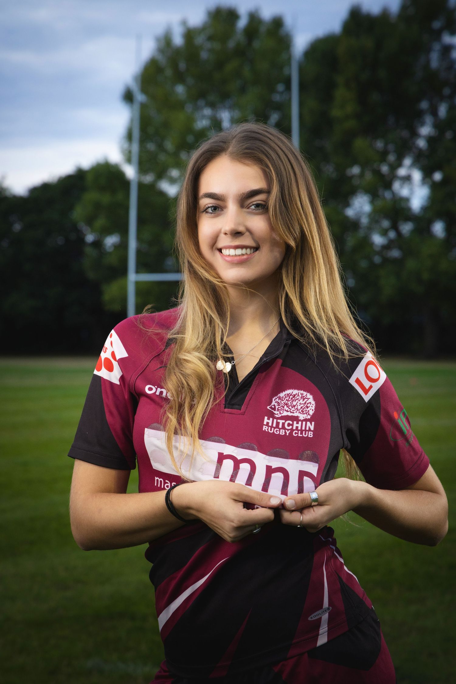 Young woman smiling in Hitchin Rugby Club maroon jersey on green field with rugby posts in background.