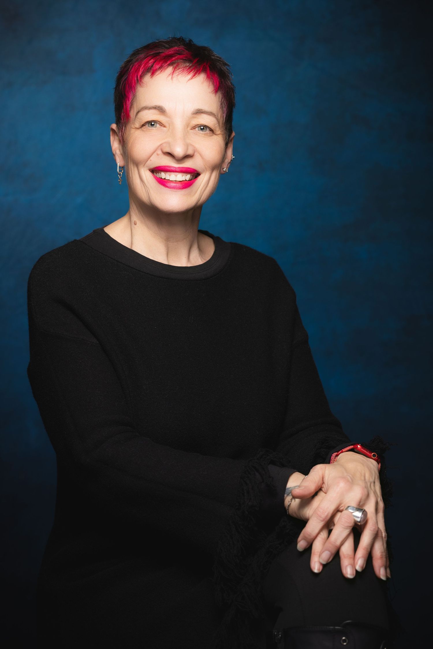 Smiling woman with short pink hair wearing black top, posing against blue background in professional portrait.
