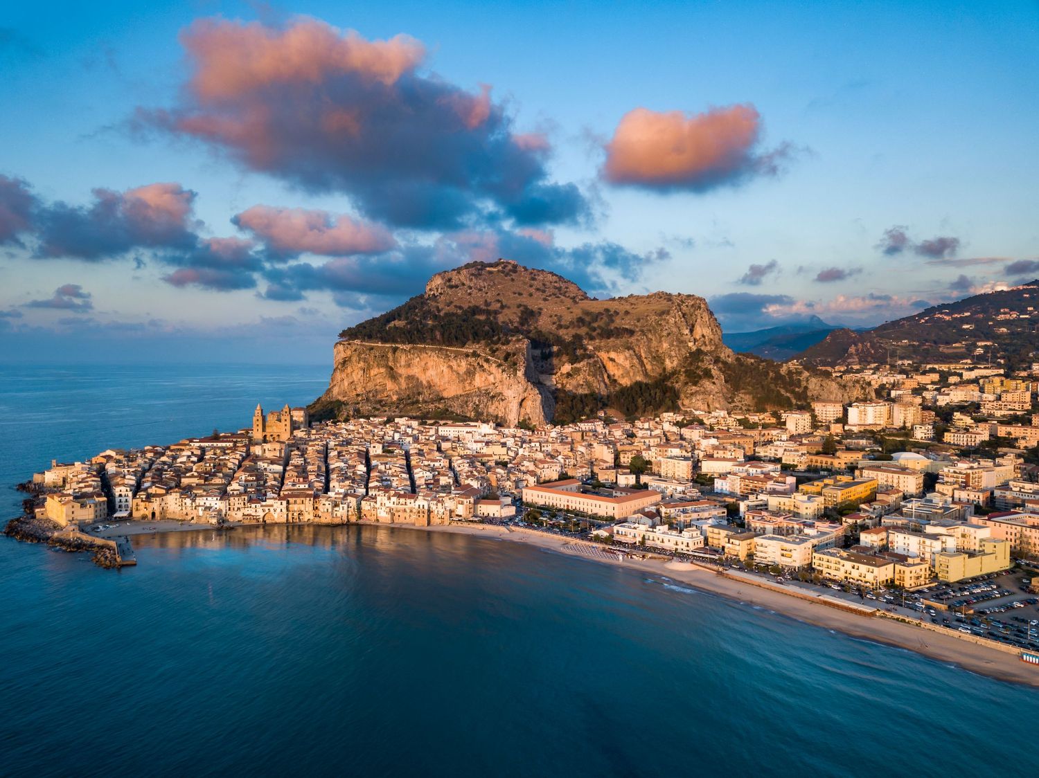 Aerial view of a picturesque coastal town nestled beneath a dramatic rocky cliff at golden hour.