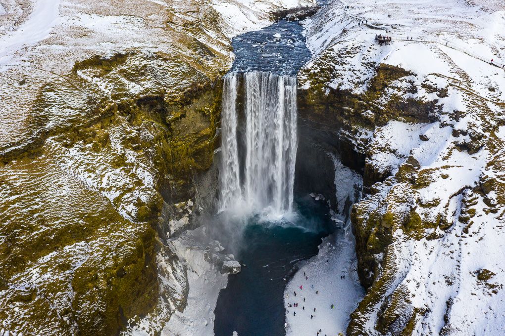 Aerial view of Skogafoss waterfall in Iceland during winter with snow covered cliffs and dark pool below.