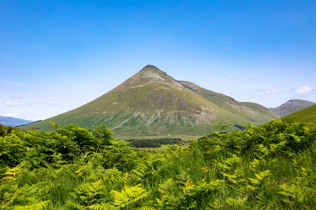 Panoramic view of green mountain peaks and valleys under a bright blue sky in Glencoe in the Scottish Highlands.