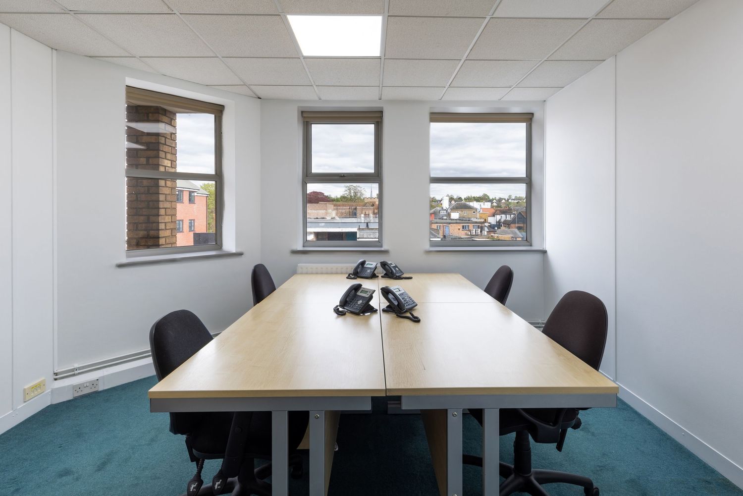 A small conference room with a long wooden table, black office chairs, two windows, and phones on a teal carpet floor.