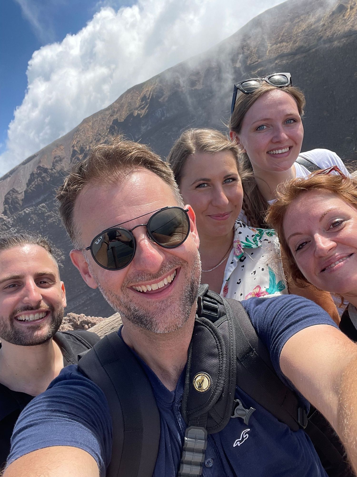 Five smiling hikers take a group selfie on a rocky volcanic slope with steam rising and white clouds in a blue sky behind them.