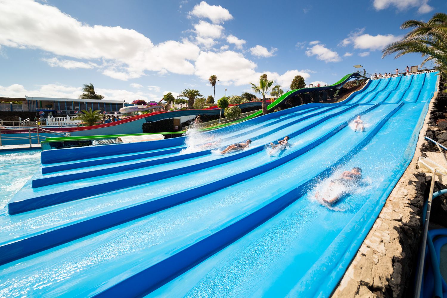 Multi-lane blue water slide at an outdoor water park with white clouds in the sky.