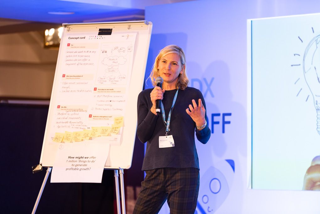 A presenter wearing a dark top gestures while speaking next to a flip chart during a conference presentation.