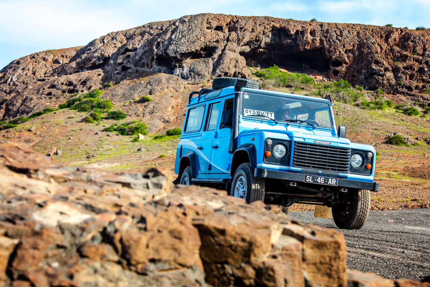 Bright blue Land Rover Defender driving along a rocky mountain road with rugged cliffs in the background.