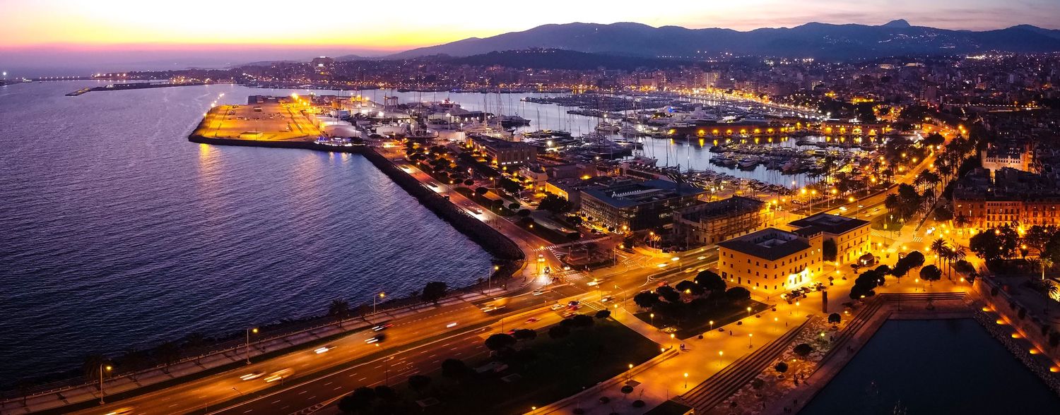 Aerial drone photograph of a coastal city harbour at dusk featuring illuminated waterfront buildings and reflections on calm water.