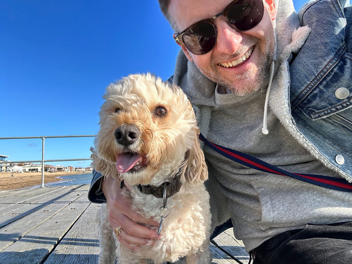 Smiling man with sunglasses posing with a fluffy golden doodle dog on a sunny boardwalk under a clear blue sky.
