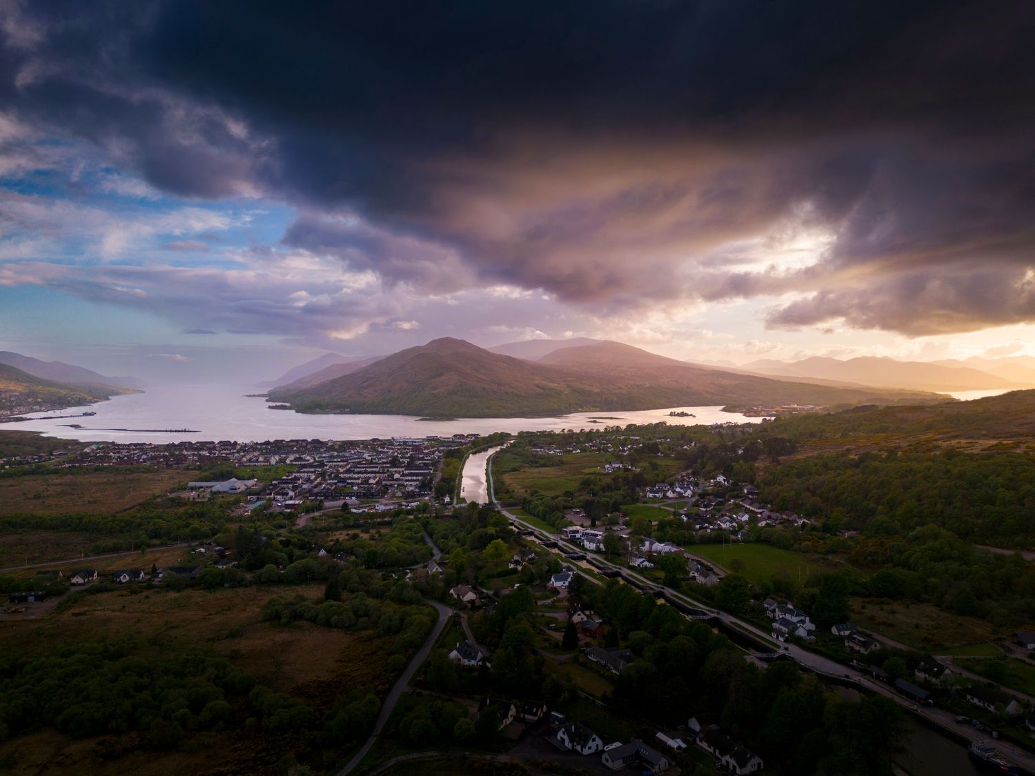 Aerial drone view of an Irish coastal town at sunset with dramatic storm clouds over mountains and a tranquil bay.