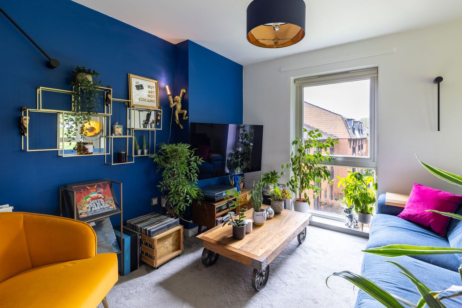 Vibrant living room with bold blue accent wall, gold shelving, TV, houseplants, mustard sofa, and wooden coffee table.