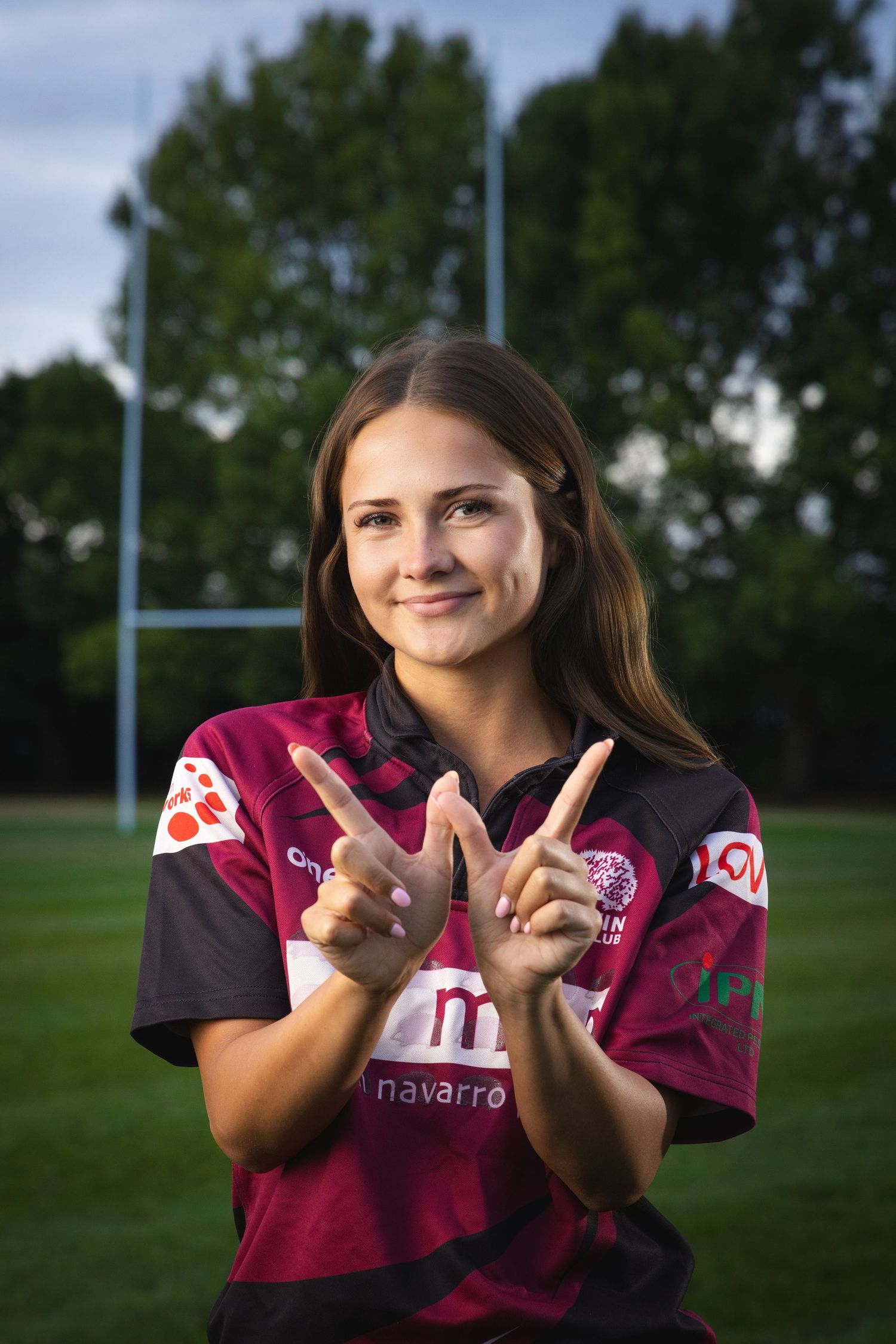 Young female rugby player in maroon jersey making hand gestures on a sports field with goalposts behind her.