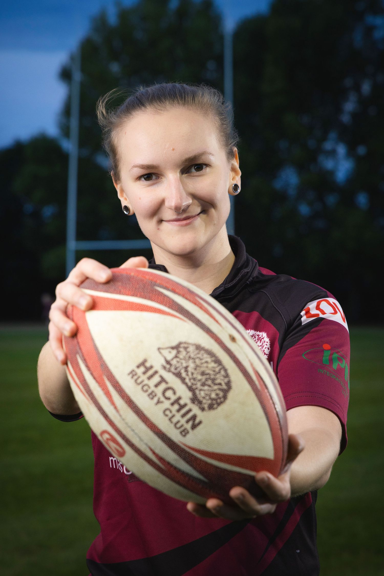 Female rugby player in maroon jersey holding a Hitchin Rugby Club ball toward camera on outdoor pitch.