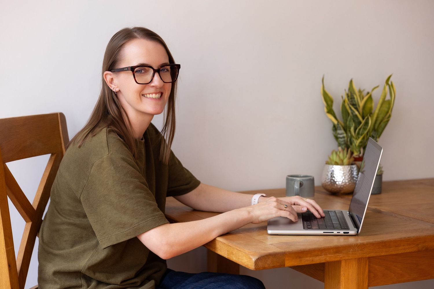 Woman smiling at camera while working on laptop at wooden desk with plant and mug nearby.