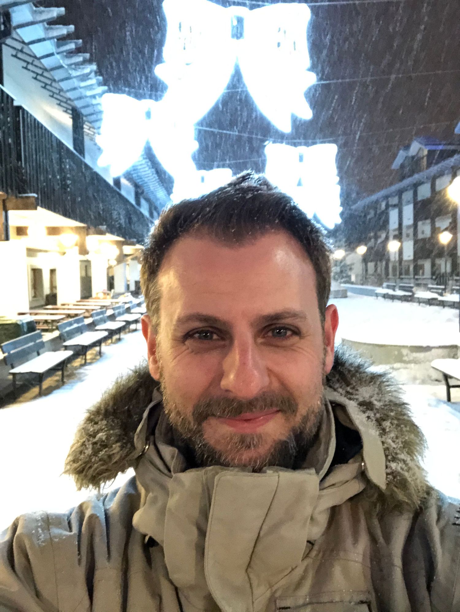 Man with beard in winter jacket smiles during snowy evening at outdoor venue with festive lights.