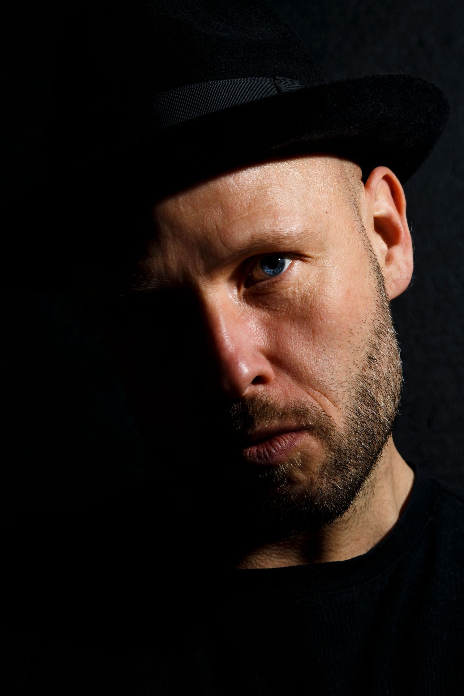 Dramatic professional portrait of man wearing a black hat, photographed with low-key lighting against a dark background.