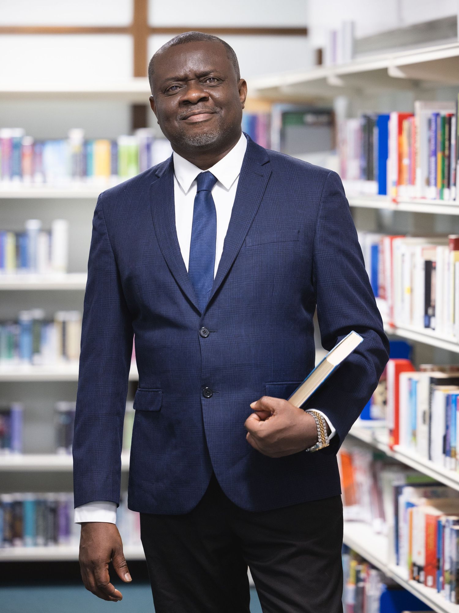 Professional man in navy suit holding books while standing in a library between colorful bookshelves.