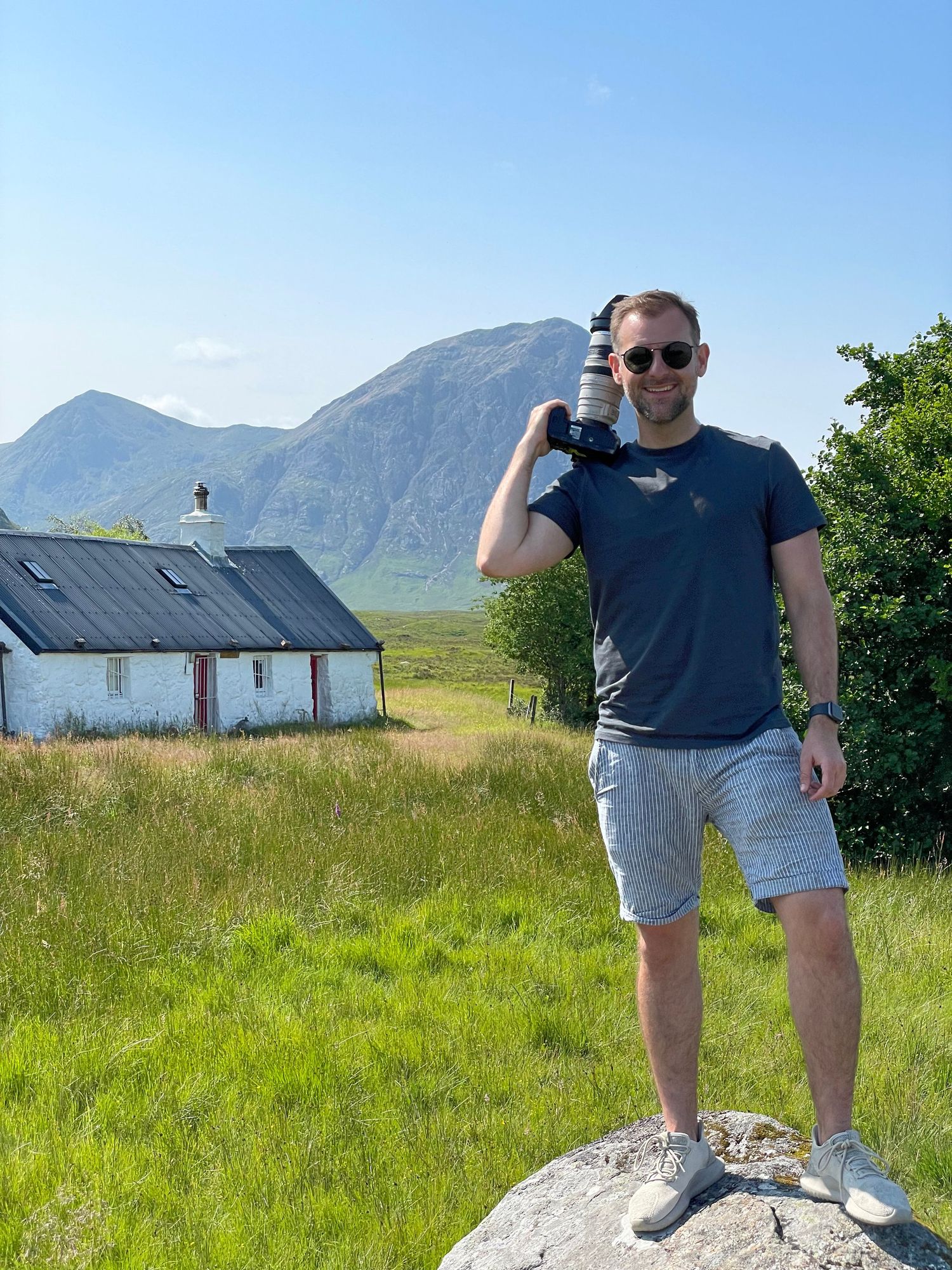 Lee Charlton photographing in the Scottish Highlands with mountain scenery and a traditional white cottage in the background.
