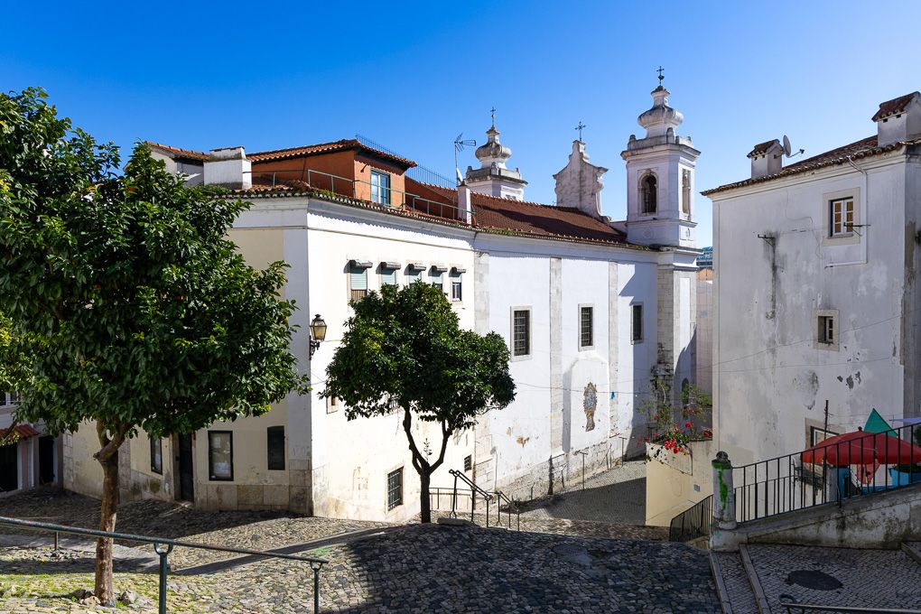 White church with twin bell towers and small courtyard surrounded by buildings in historic Lisbon, Portugal.