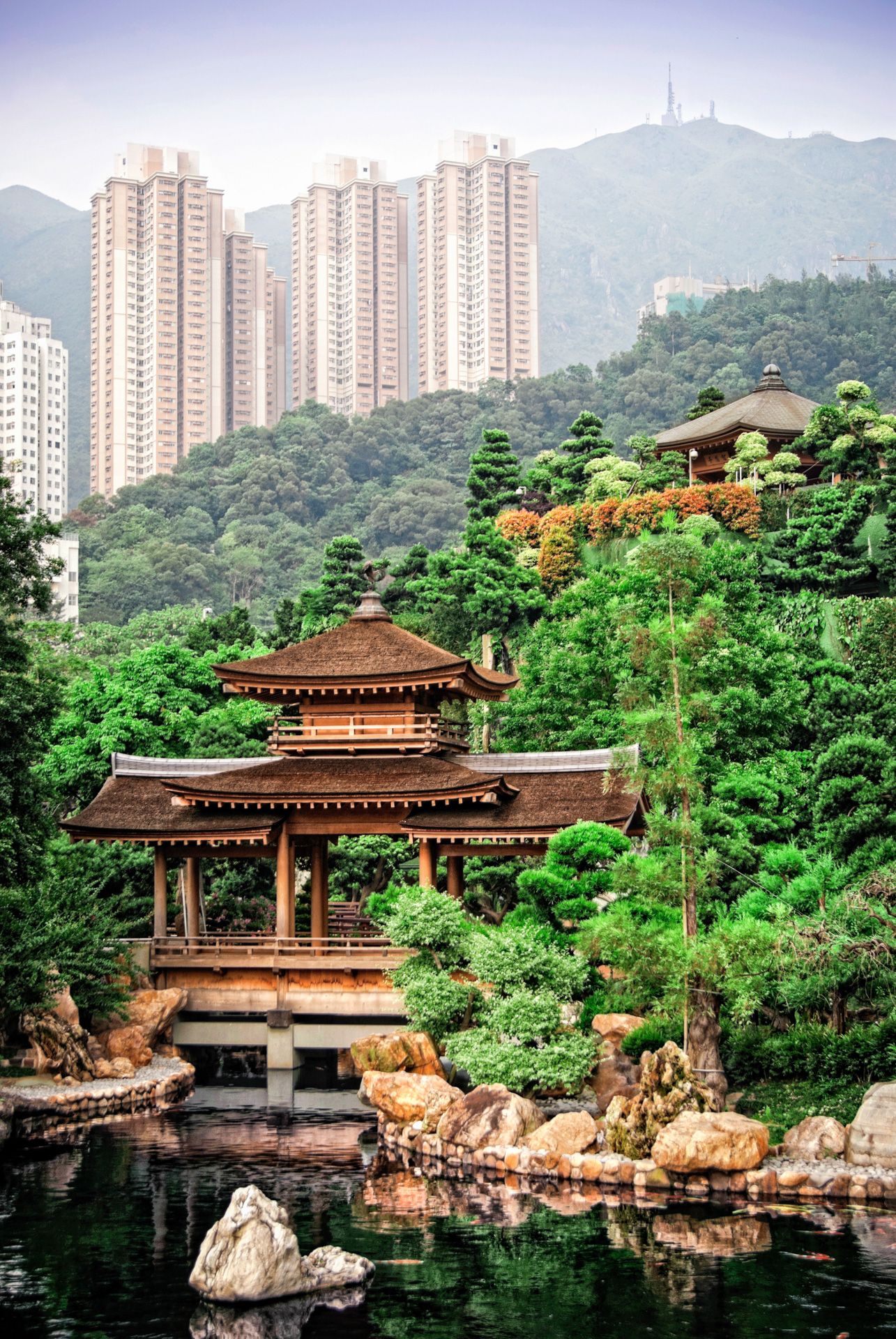 Traditional Chinese pagoda pavilion with multiple tiers overlooks a tranquil pond against backdrop of high-rise buildings and mountains.