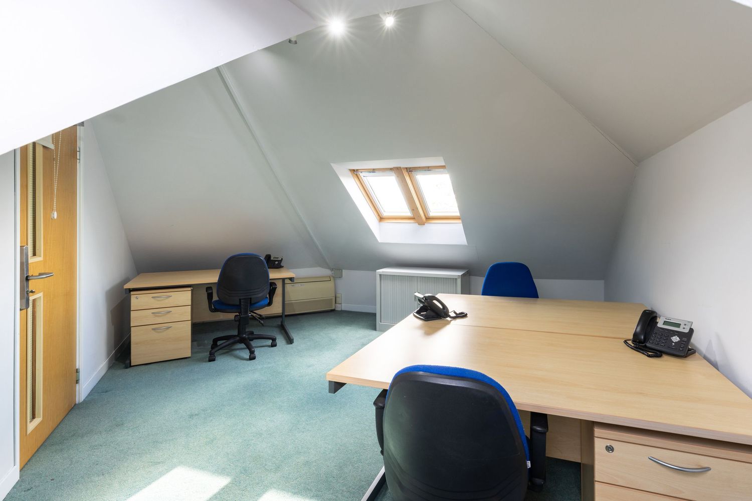 Bright attic office space with sloped ceilings, skylight, wooden desks, blue chairs, and teal carpet flooring.