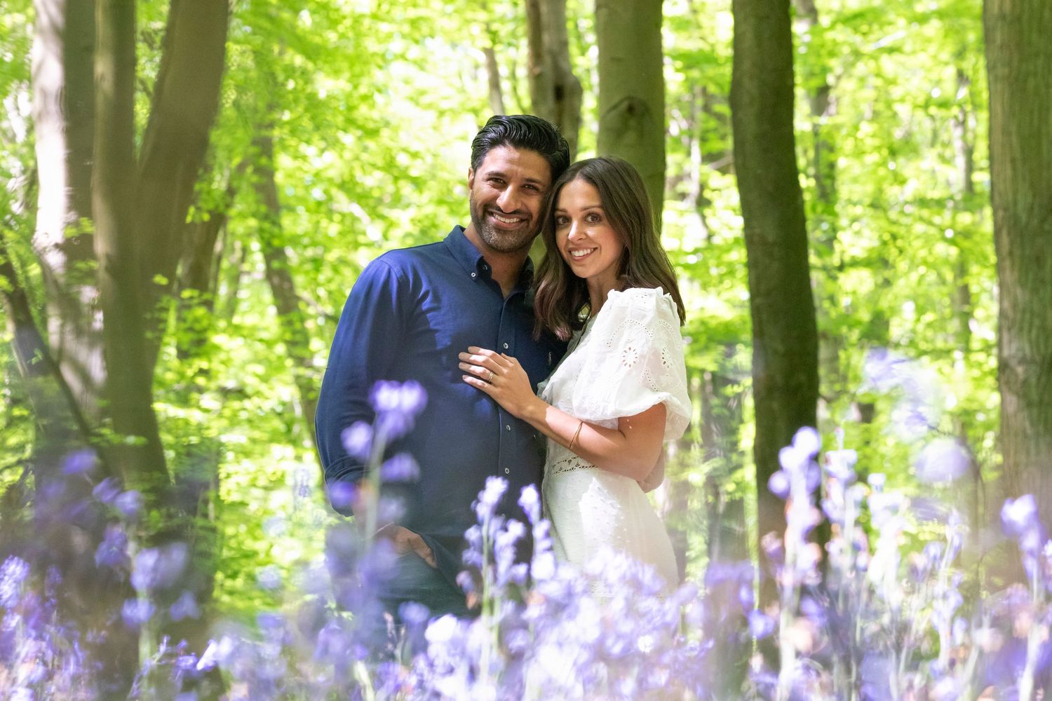 Couple smiling together in a bluebell woodland forest, surrounded by purple flowers and lush green trees.