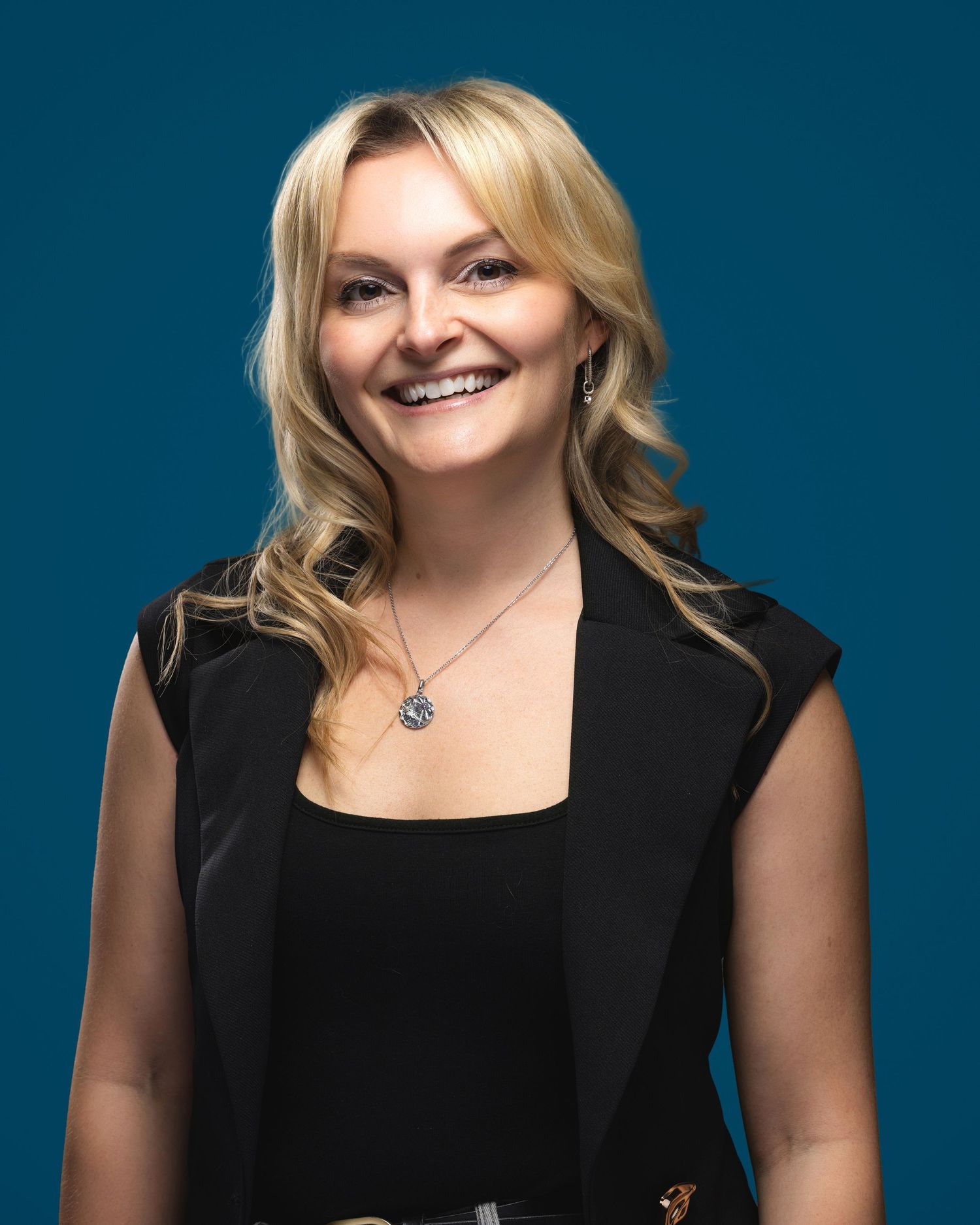 Professional woman with blonde hair smiling in black blazer against teal background.
