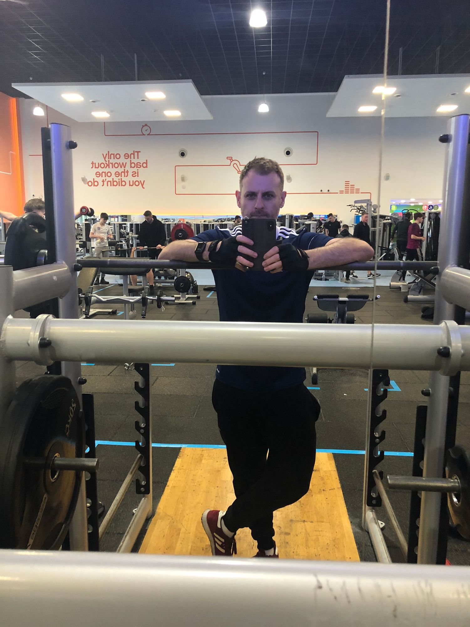 Man taking a mirror selfie at the gym, showing muscular arms and wearing black workout clothes.