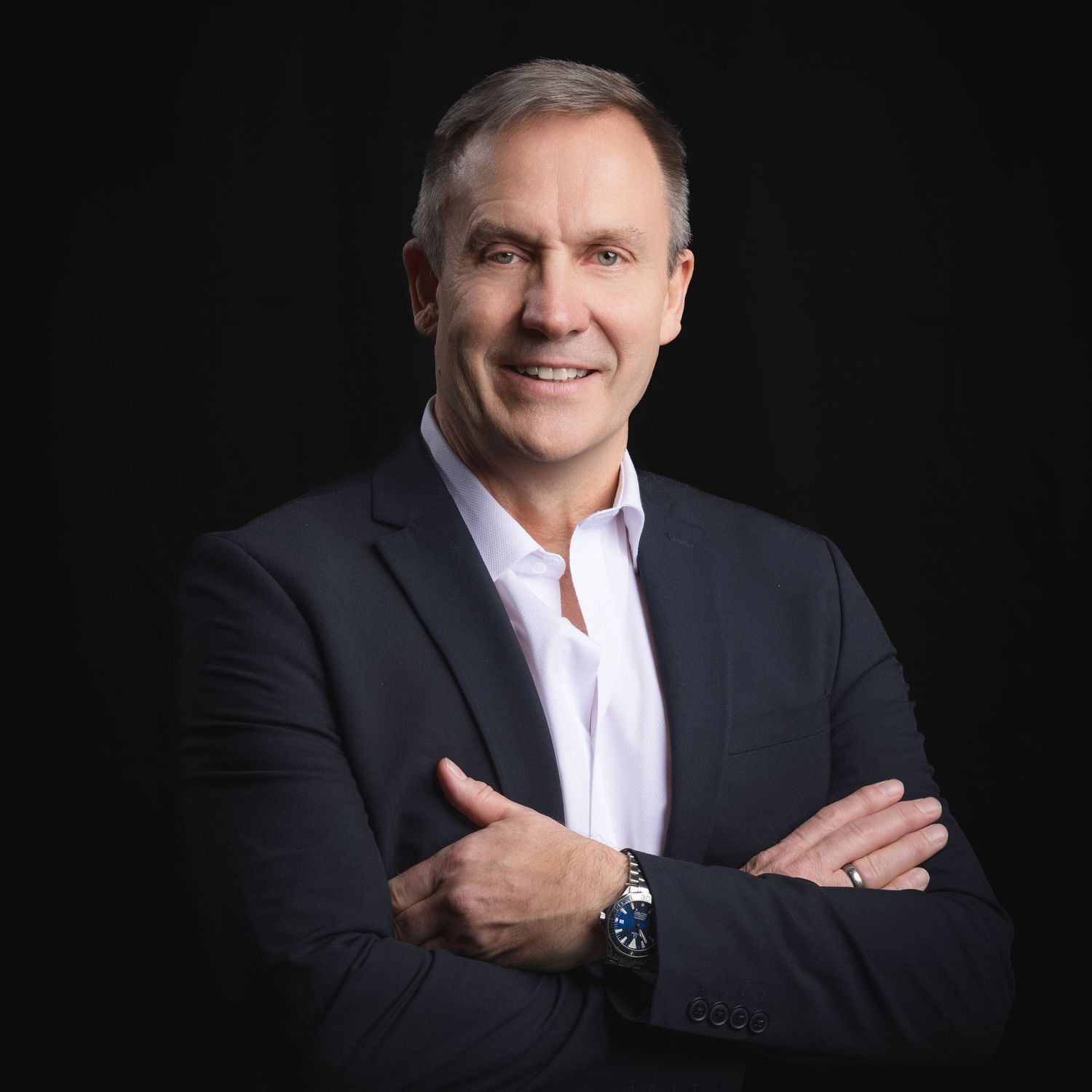 Professional headshot of a smiling middle-aged man in a navy blazer with arms crossed against a dark background.