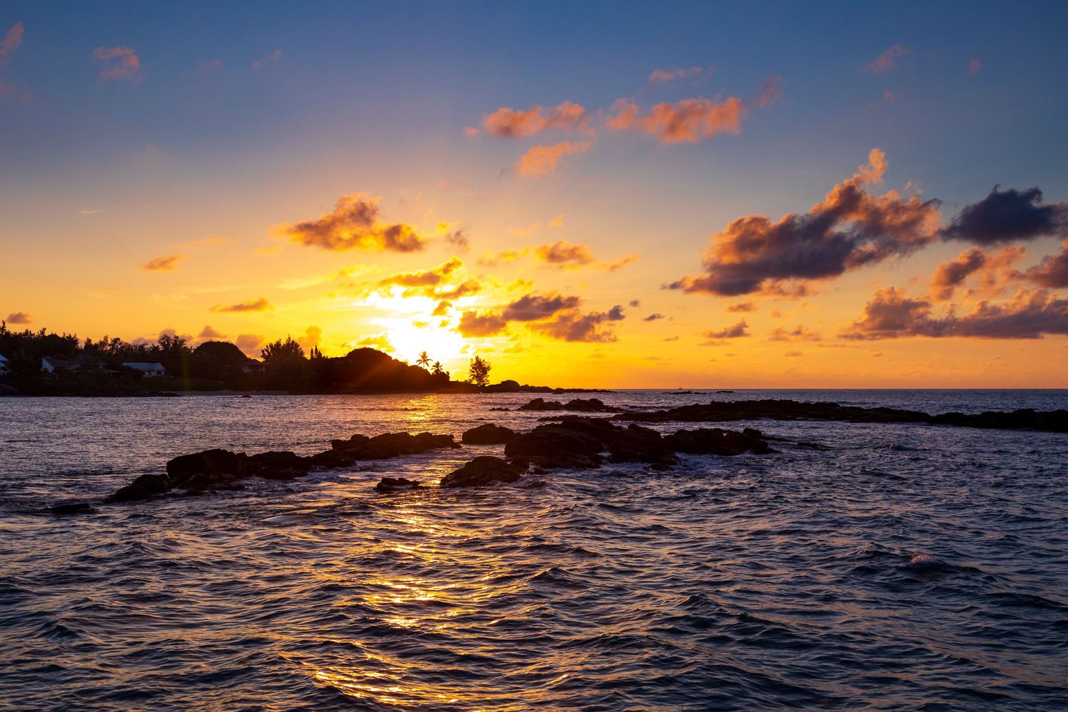 Stunning ocean sunset with golden sky, rocky shoreline silhouette, and calm waters in a tropical coastal setting.