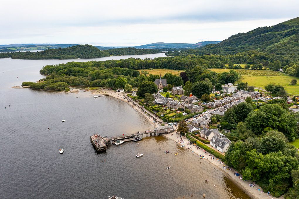 Aerial panoramic view of Luss on Loch Lomand, with boats anchored in the bay and lush green hills surrounding the shoreline.